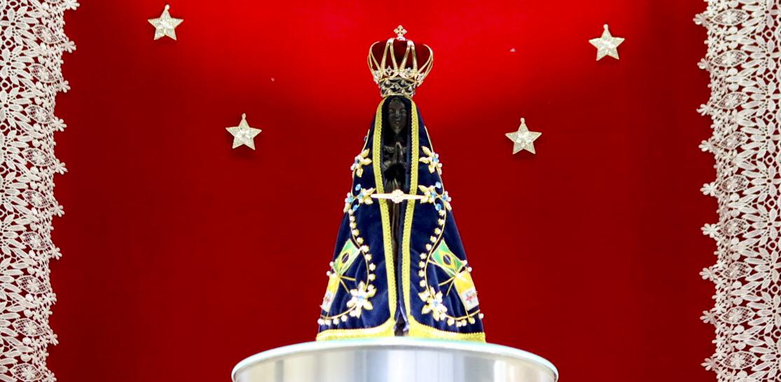 em primeiro plano imagem de Nossa Senhora Aparecida vestida com manto azul e coroa dourada sobre Altar circular prateado,; Ao fundo tecido vermelho aveludado com estrelas douradas. 